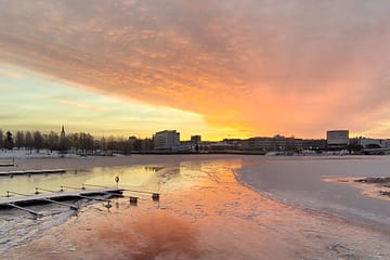 kuva auringonlaskusta pakkasella, kuvassa tyhjiä venelaitureita ja taustalla Oulun teatteri ja kirjasto, Kuvattu raatin vierasvenesatamassa.